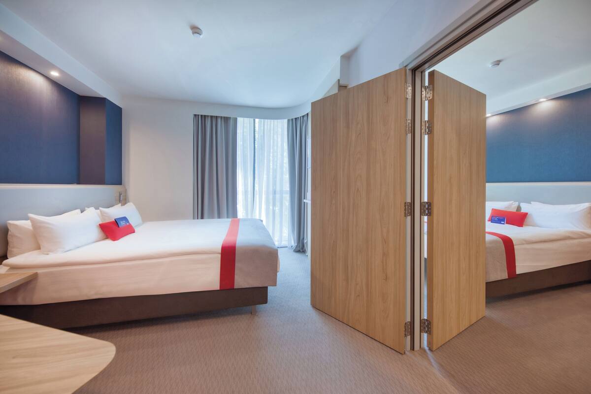 A modern hotel room with two beds, a wooden wardrobe, and a large window with curtains.