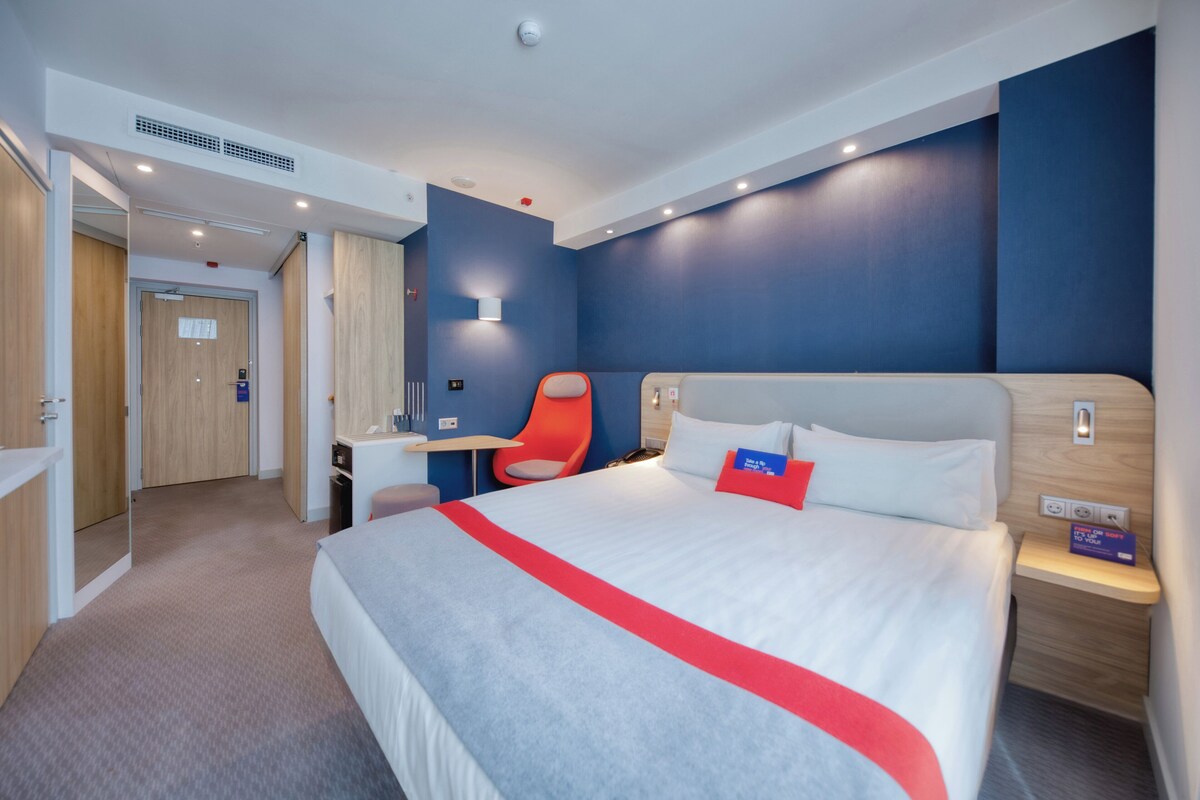 Una habitación de hotel moderna con una cama grande, un escritorio, una silla y un baño visible a través de una puerta abierta.