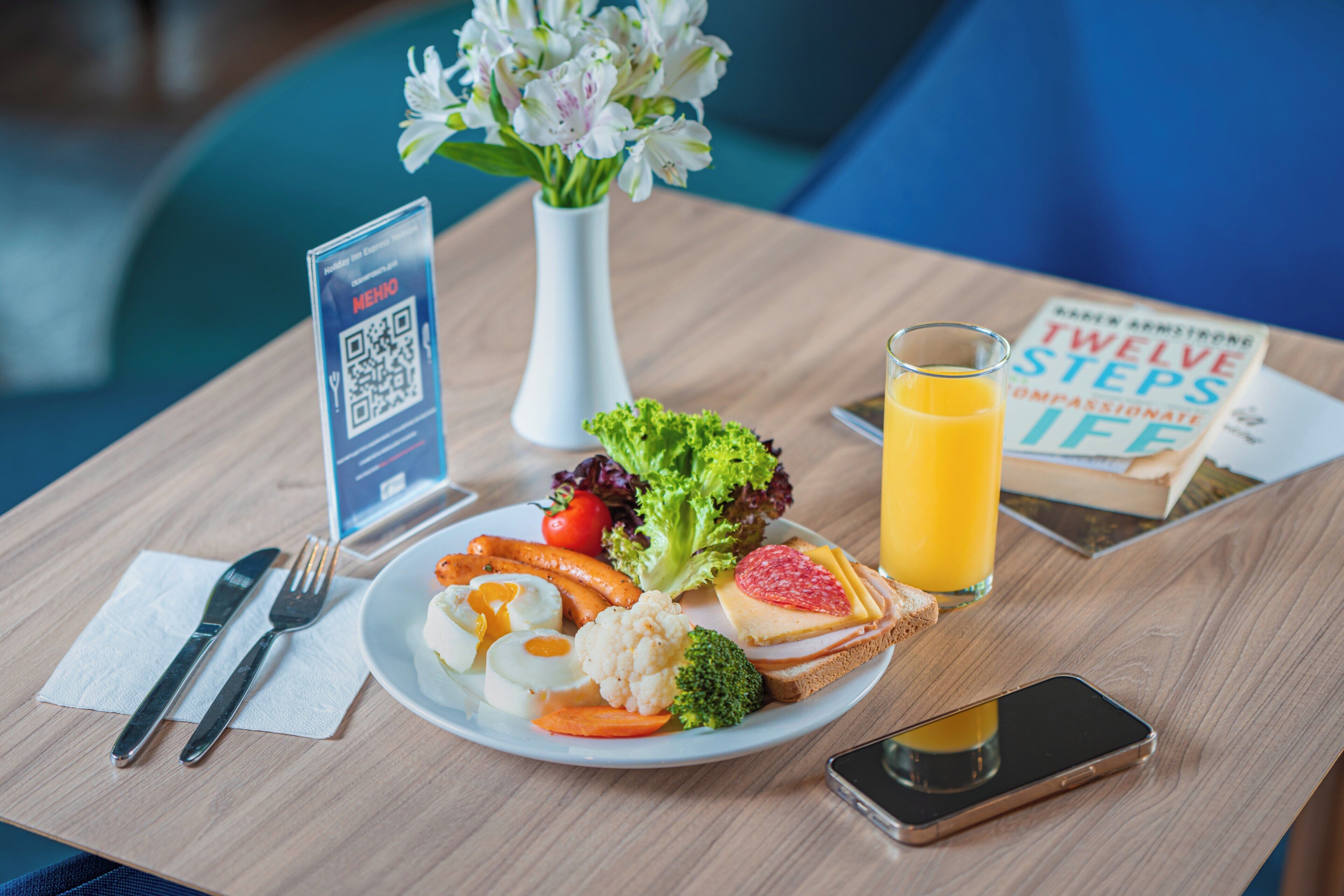 En måltid bestående av ett glas apelsinjuice, en bok, en smartphone och en skylt med en QR-kod, placerat på ett träbord.