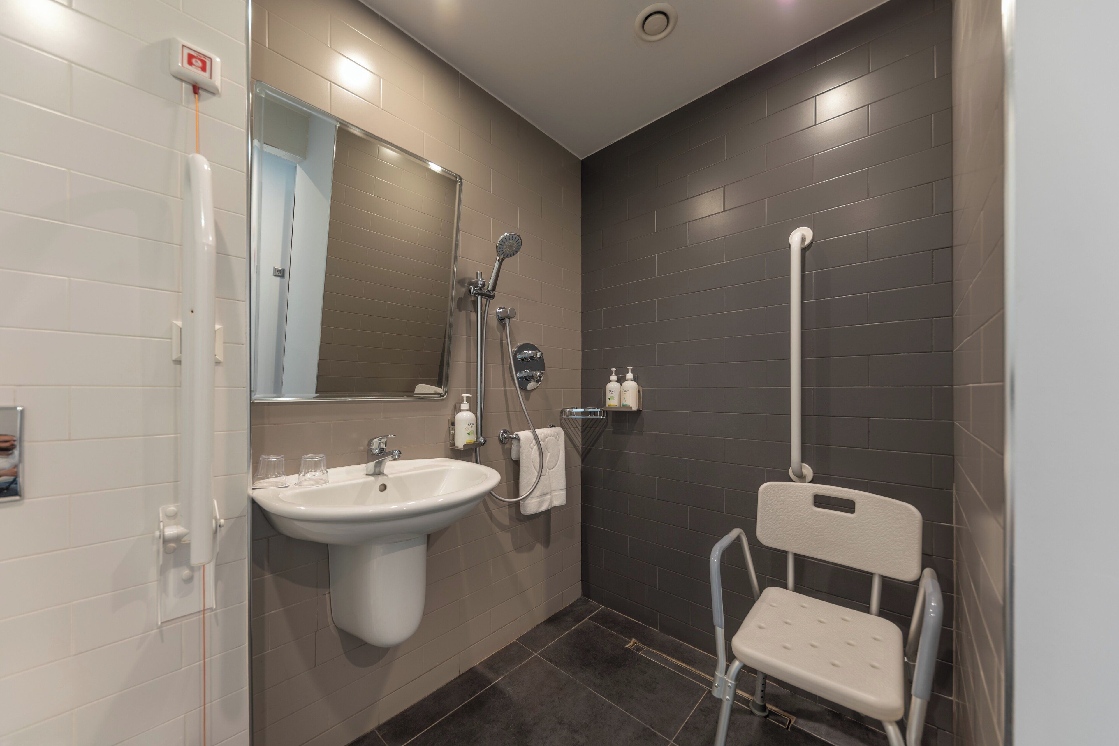 A modern bathroom with a sink, mirror, shower, and a wheelchair-accessible chair.