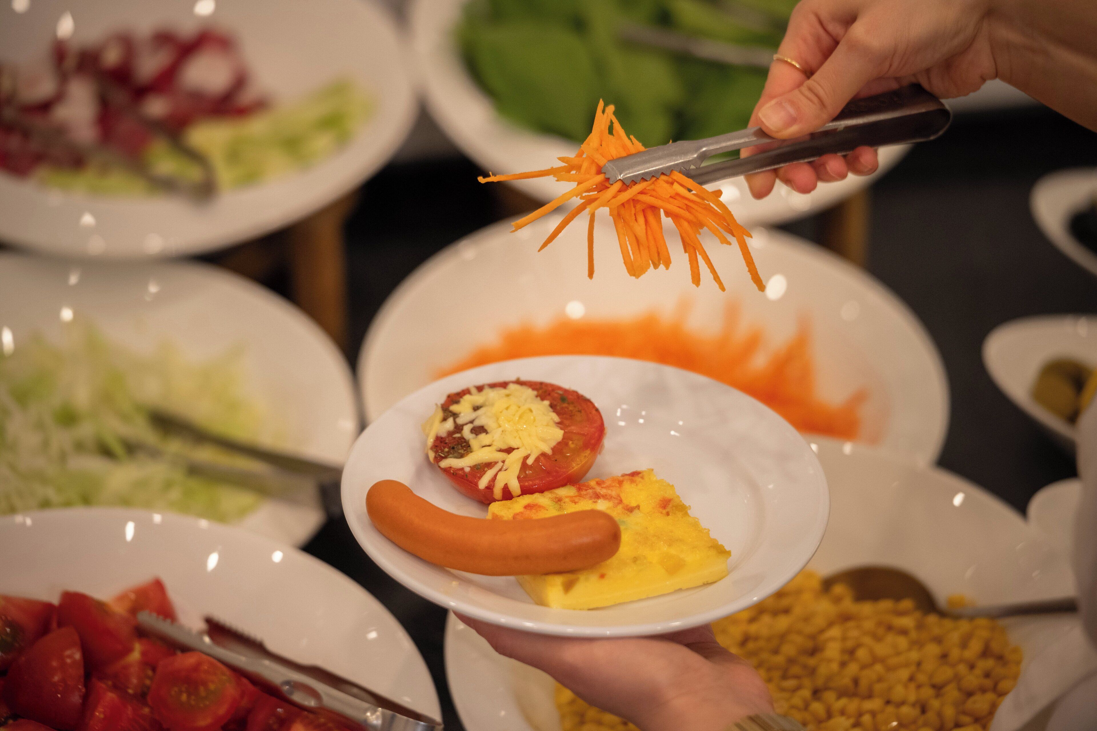 Una persona sosteniendo un plato con un perro caliente y una rodaja de tomate, rodeado de diferentes alimentos.