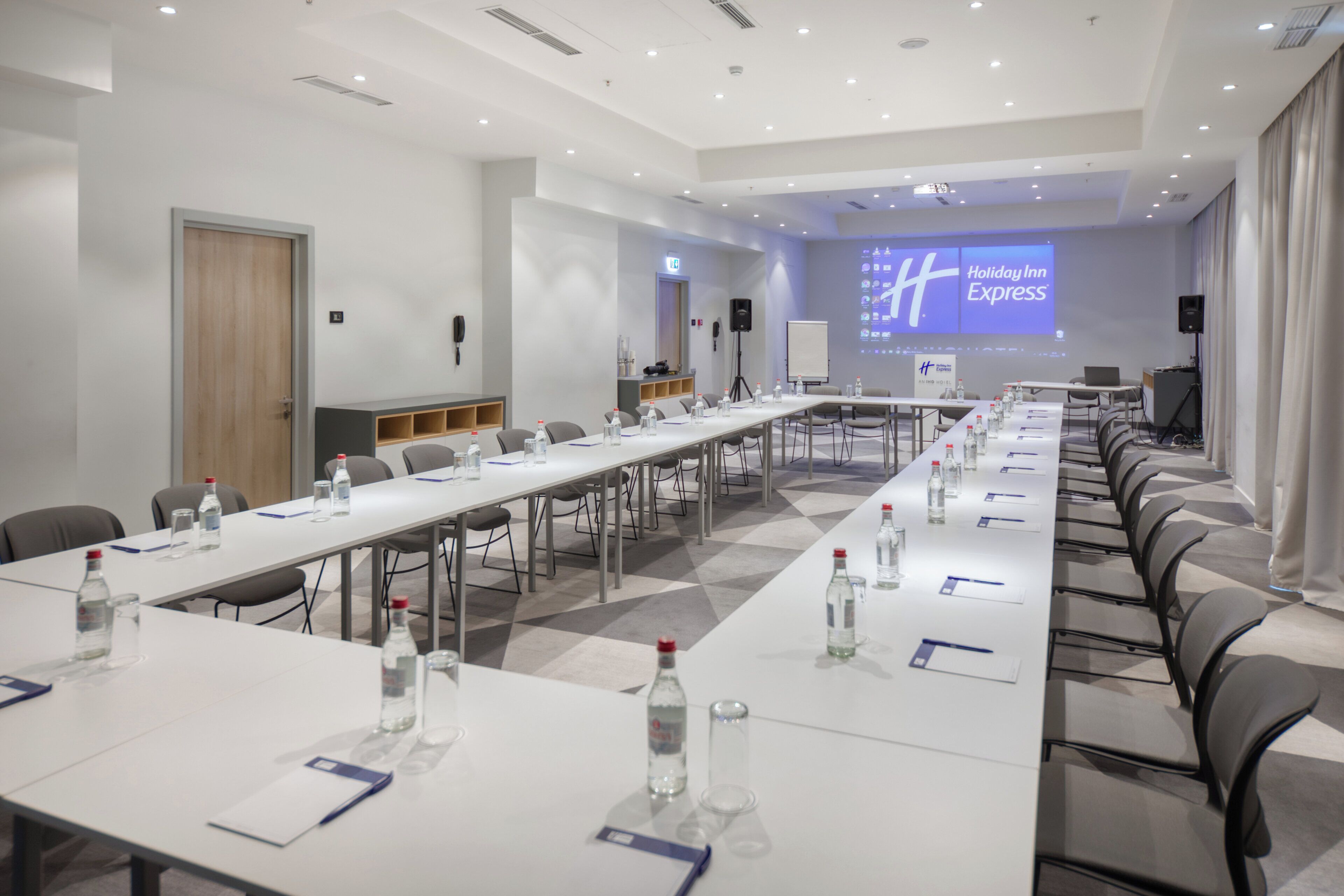 A conference room with a long table, chairs, water bottles, and a projector screen displaying the Holiday Inn Express logo.