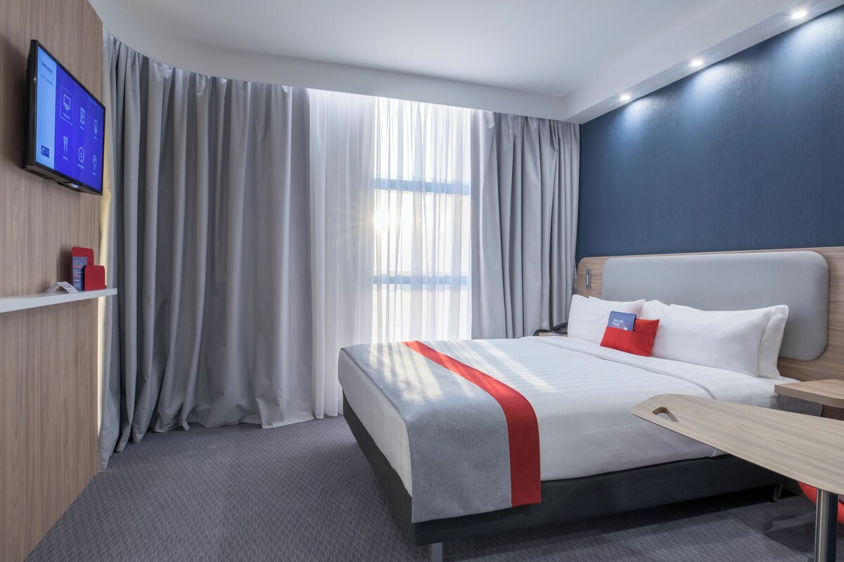 Una habitación de hotel moderna con una cama grande, un televisor de pantalla plana montado en la pared y una ventana con cortinas traslúcidas.