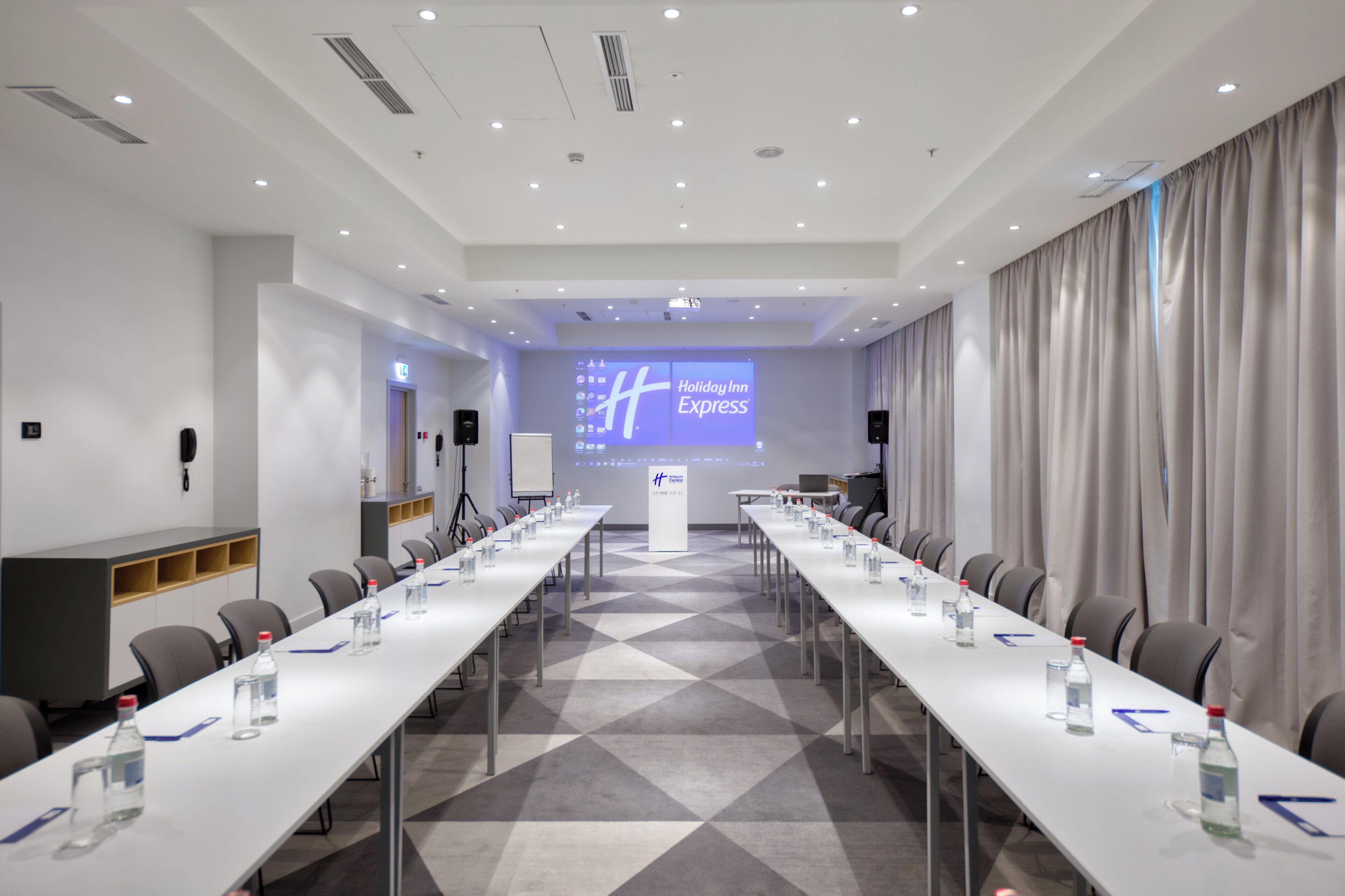 Ein Konferenzraum mit einem langen Tisch, Stühlen, einem Rednerpult und einem Bildschirm, der 'Holiday Inn Express' anzeigt.