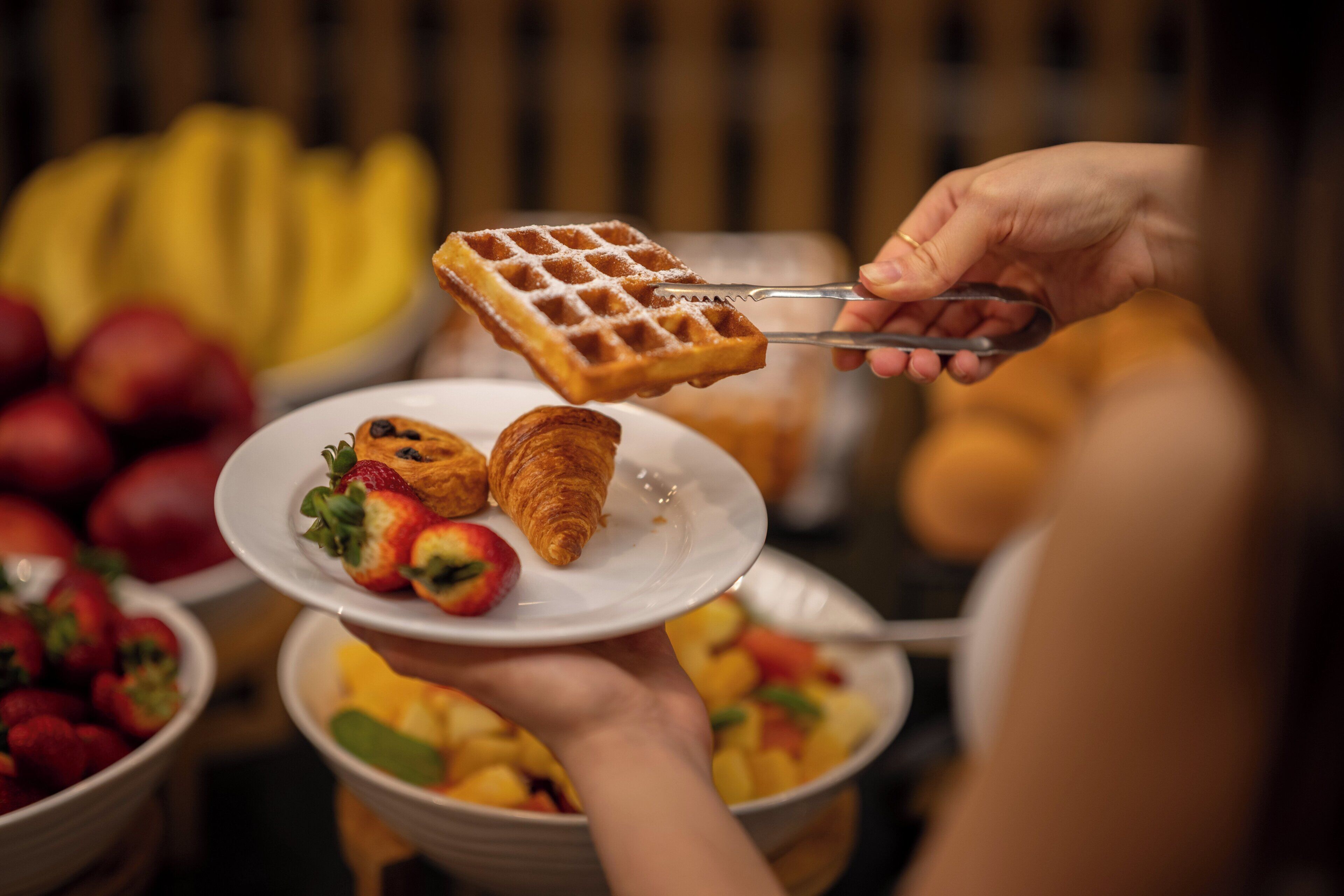 Gofre que se serveix amb pinces, acompanyat d'un croissant i maduixes sobre un plat.