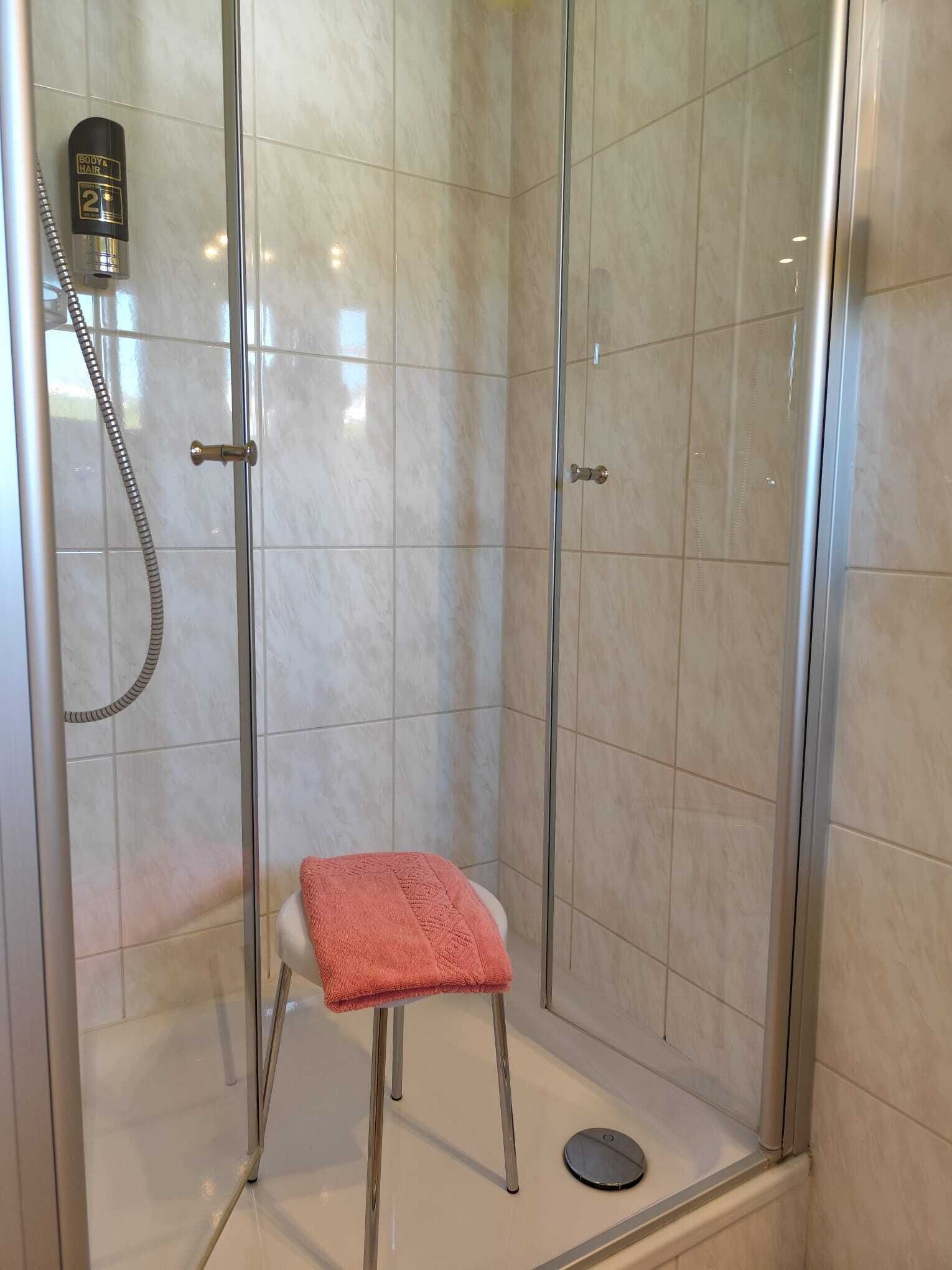 A shower area with a glass door, a handheld showerhead, and a small stool with a towel.
