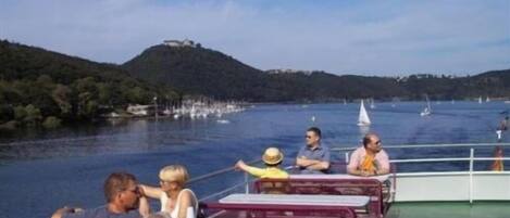 Un bote con varias personas sentadas en mesas, una vista de un lago con barcos y una colina al fondo.