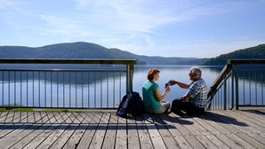 Two people sitting on a wooden dock overlooking a lake with hills in the background. - Garni Hotel & Ferienwohnungen Seeschlößchen (Waldeck)