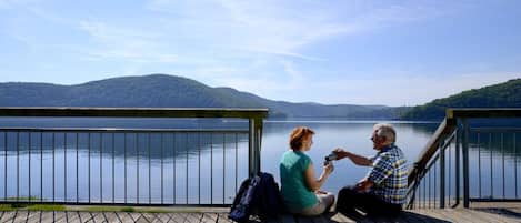 Two people sitting on a wooden dock overlooking a lake with hills in the background.