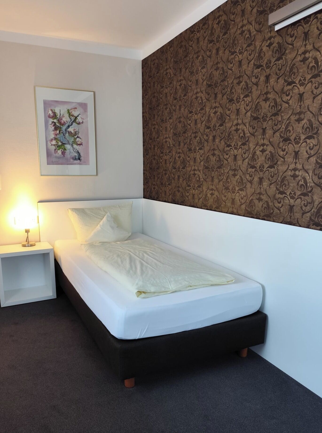 A neatly made bed with a white mattress and dark wooden frame, a small bedside table with a lamp, and a patterned wallpaper on the wall.