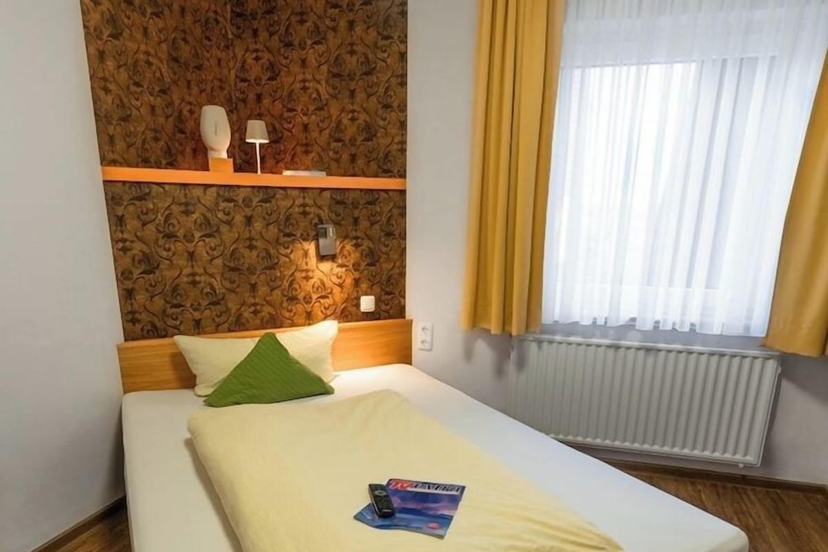 A neatly made bed with a green pillow, a bedside table with a lamp, and a shelf with decorative items.