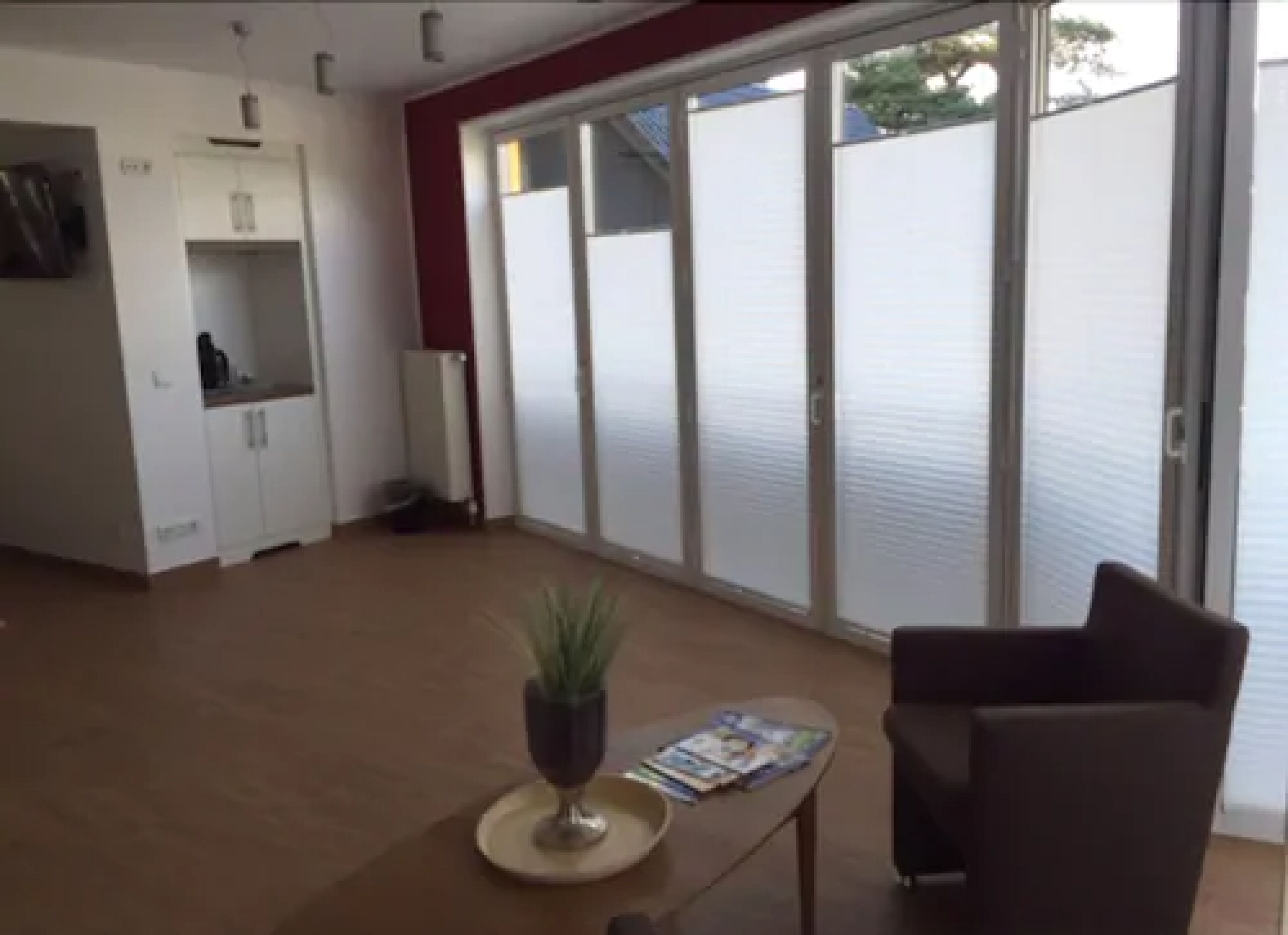 A spacious living area with a sliding glass door leading to a balcony, a dark armchair, a small table with a plant, and a white cabinet with a microwave.