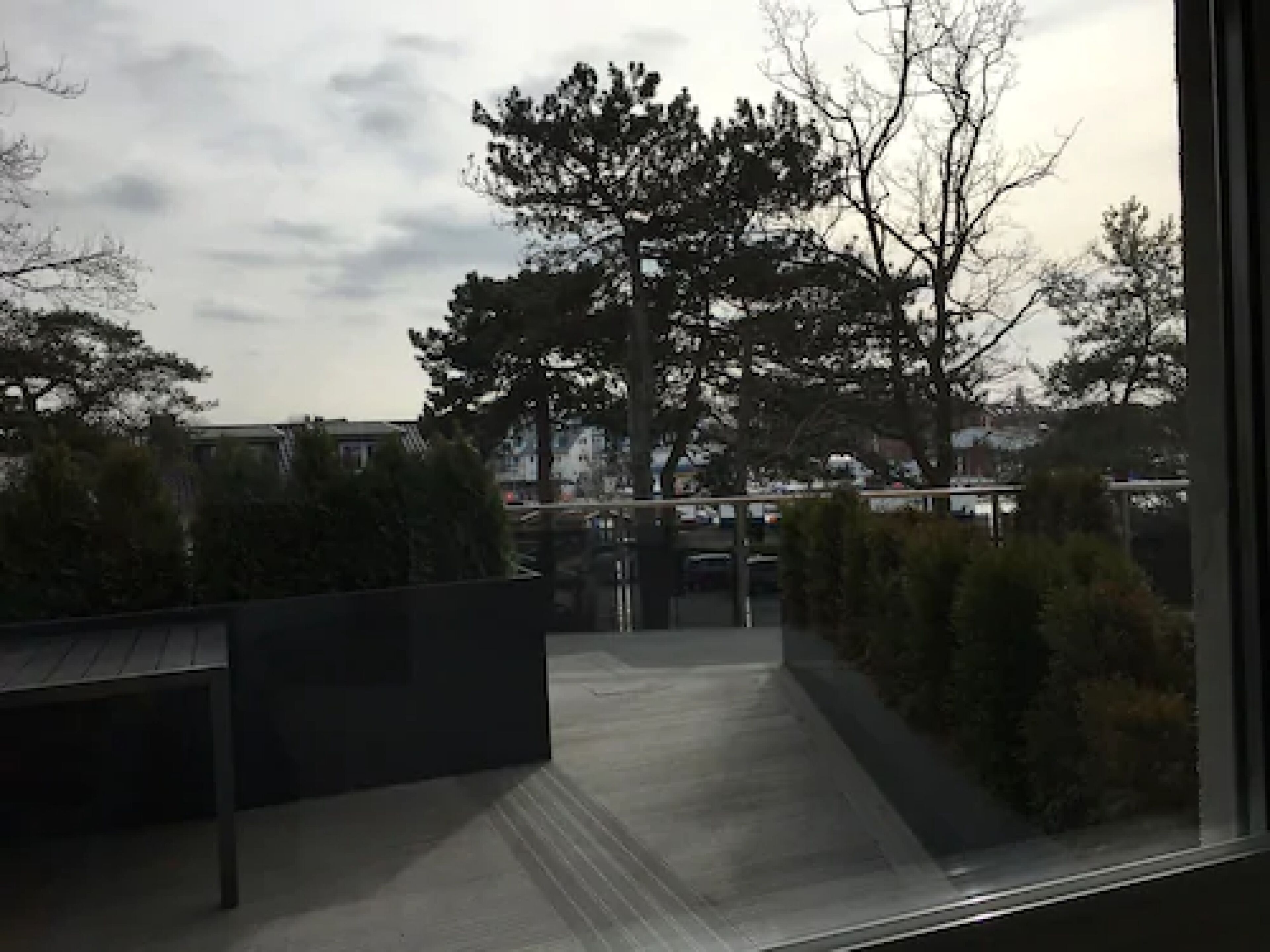 A view through a window of a building with a paved pathway leading to a bridge, surrounded by trees and buildings in the background.