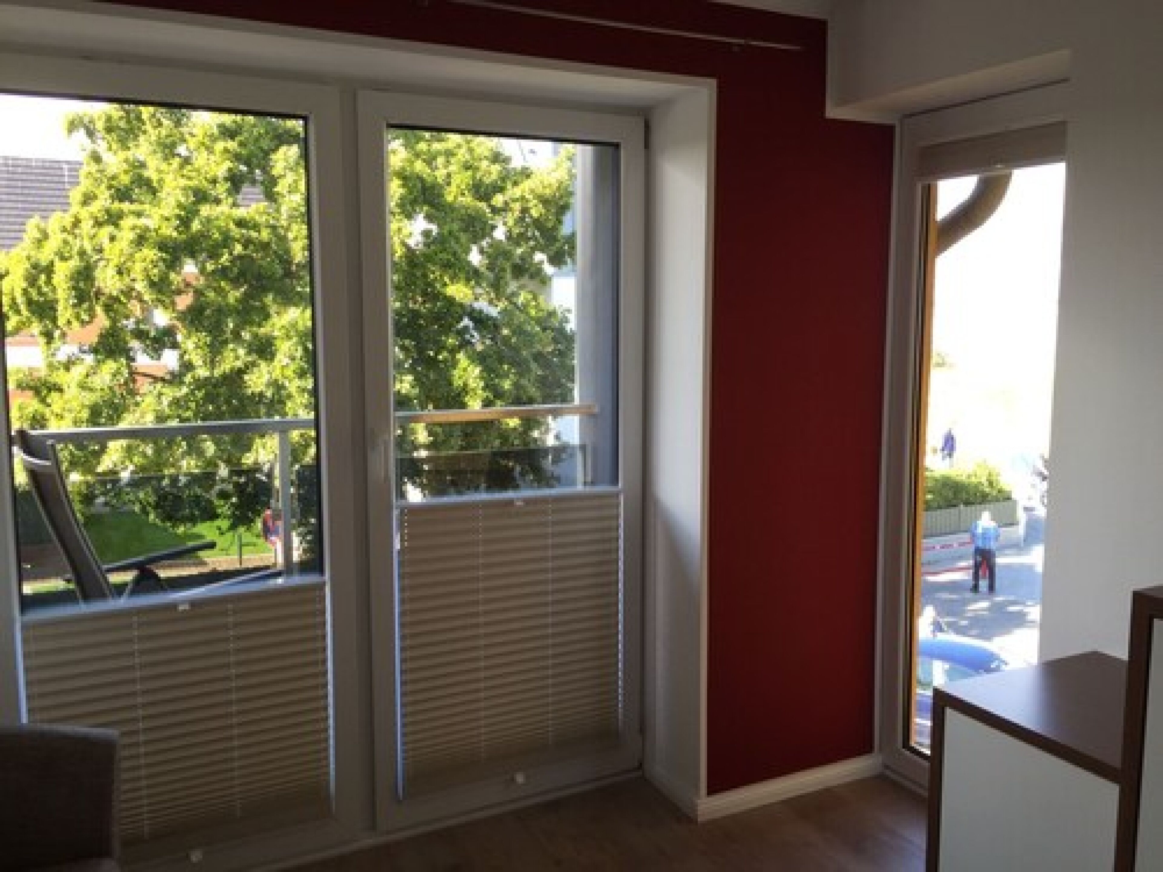 Een kamer met grote schuifdeuren naar een balkon, een rode accentmuur en uitzicht op een woonwijk met bomen en geparkeerde auto's.