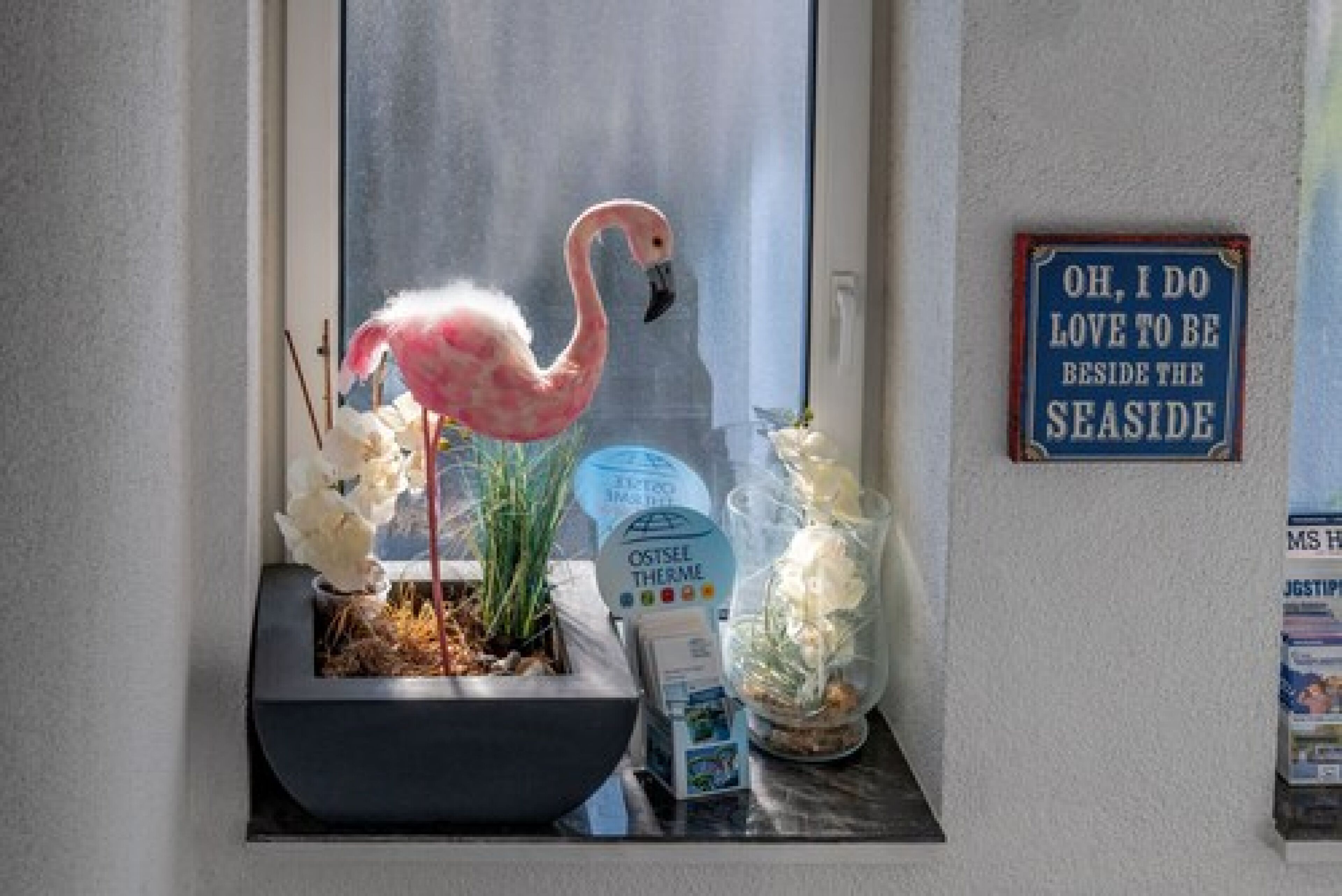 Ein Fenster, dekoriert mit einem Flamingo, einem Schild mit der Aufschrift "OH, I DO LOVE TO BE BESIDE THE SEASIDE" und einer kleinen Pflanze.