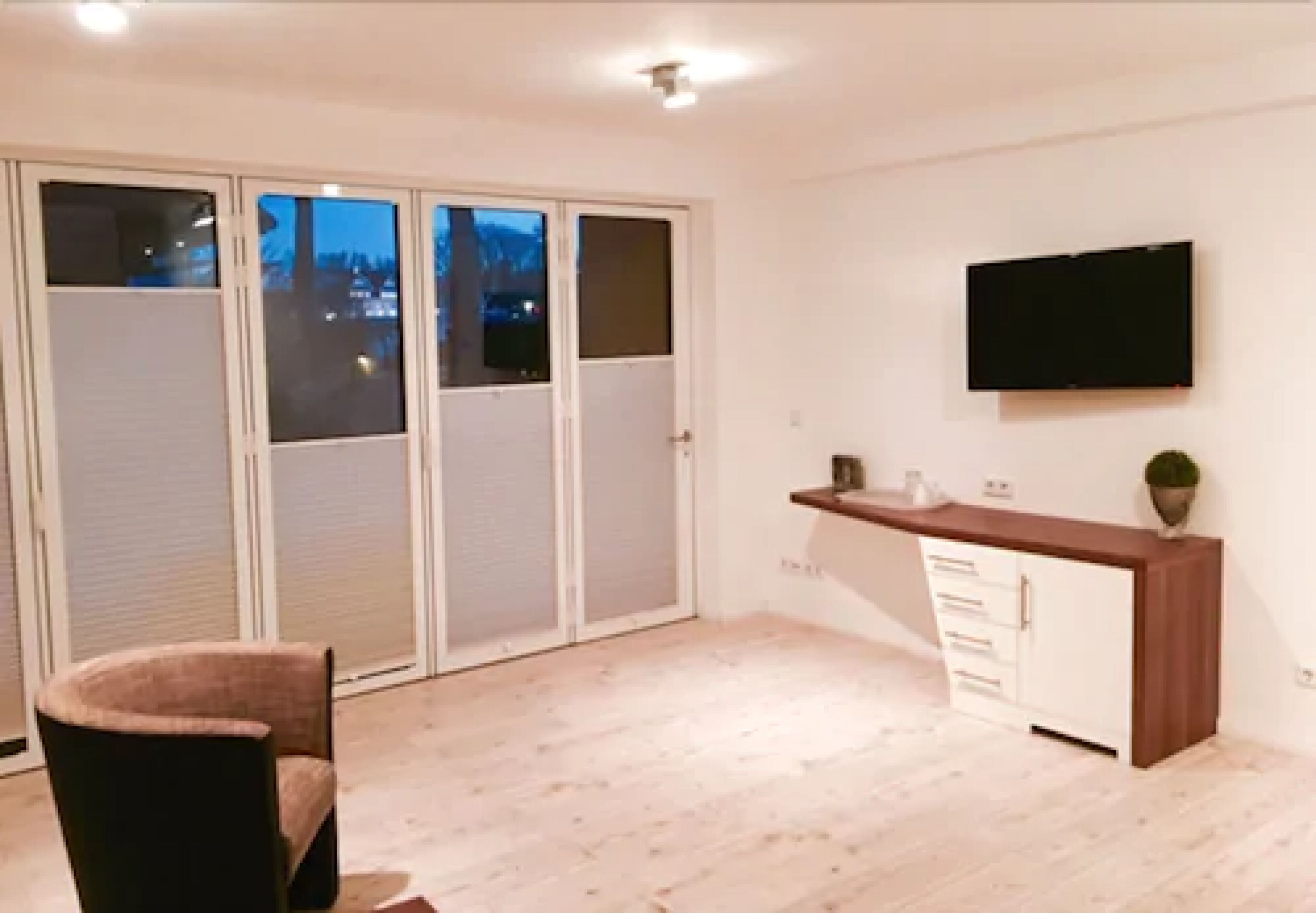 Habitació amb porta corredissa de vidre, televisió pantalla plana penjada a la paret, moble de fusta i una cadira.