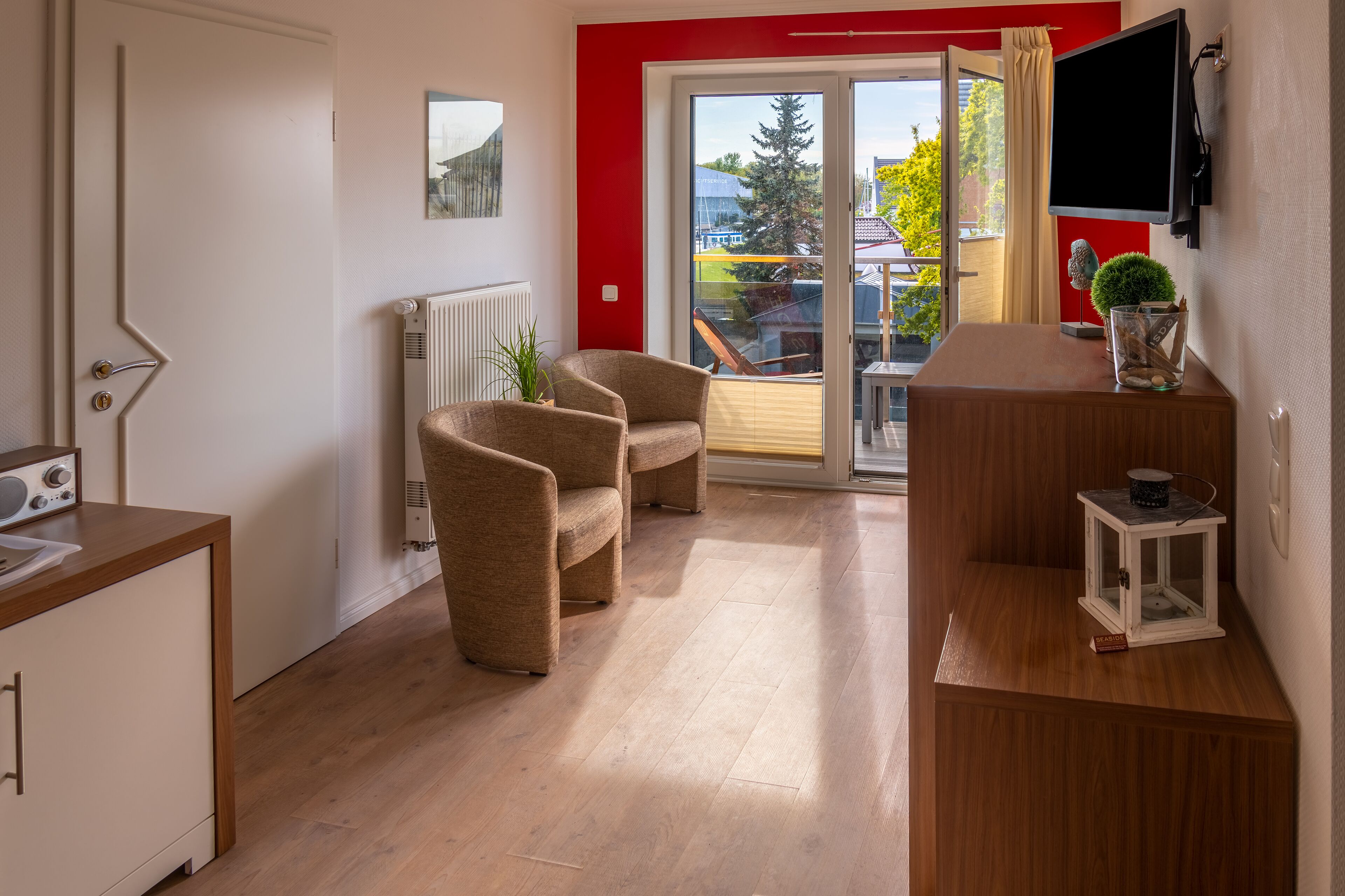Una sala de estar moderna con suelo de madera, dos sillones, una mesita, un televisor montado en la pared y un ventanal corredero que da a un balcón.