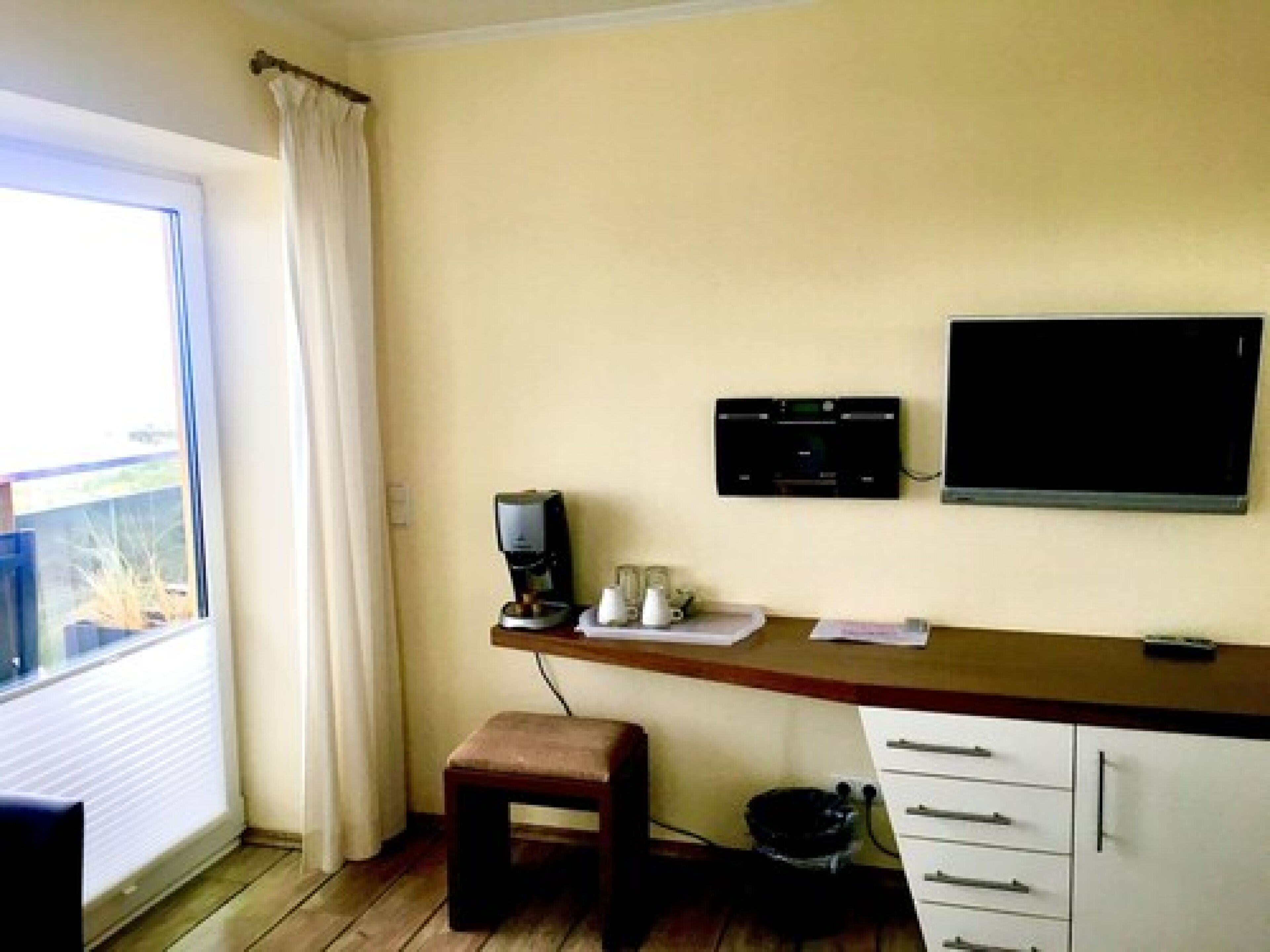 A small room with a desk, a coffee machine, a TV, and a window with curtains.