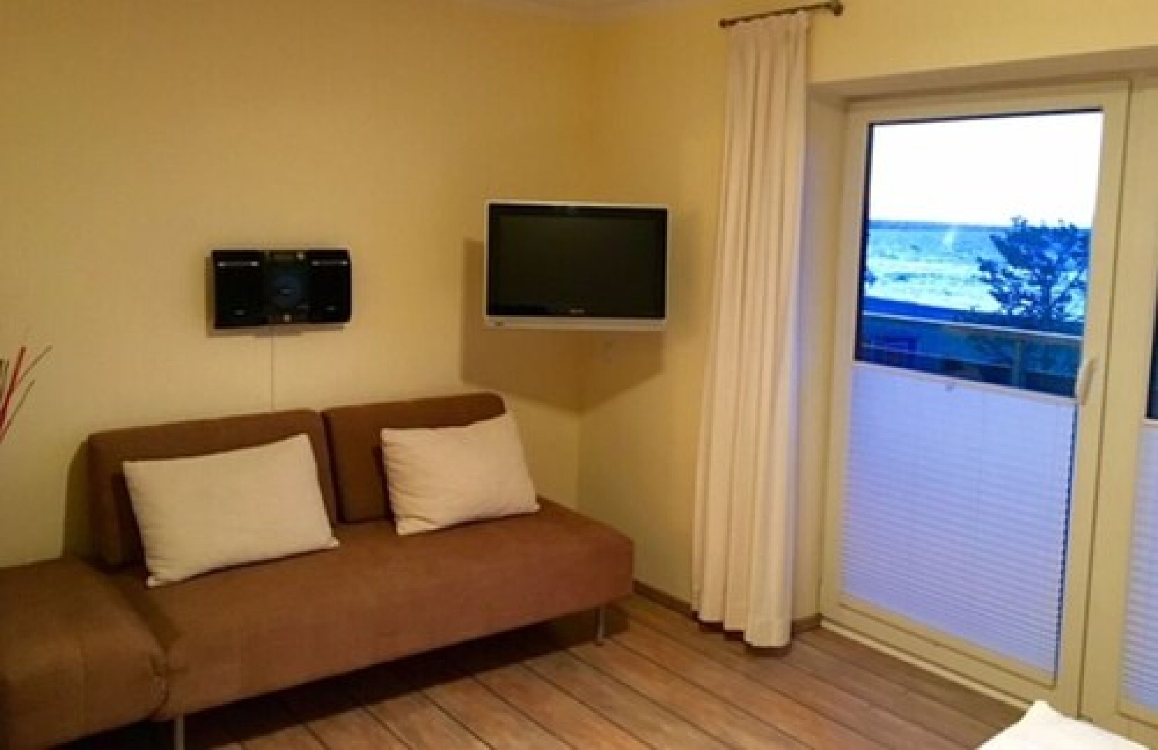 Habitación con sofá marrón, televisor de pantalla plana y puerta corrediza de vidrio que da a un balcón con vista al mar.