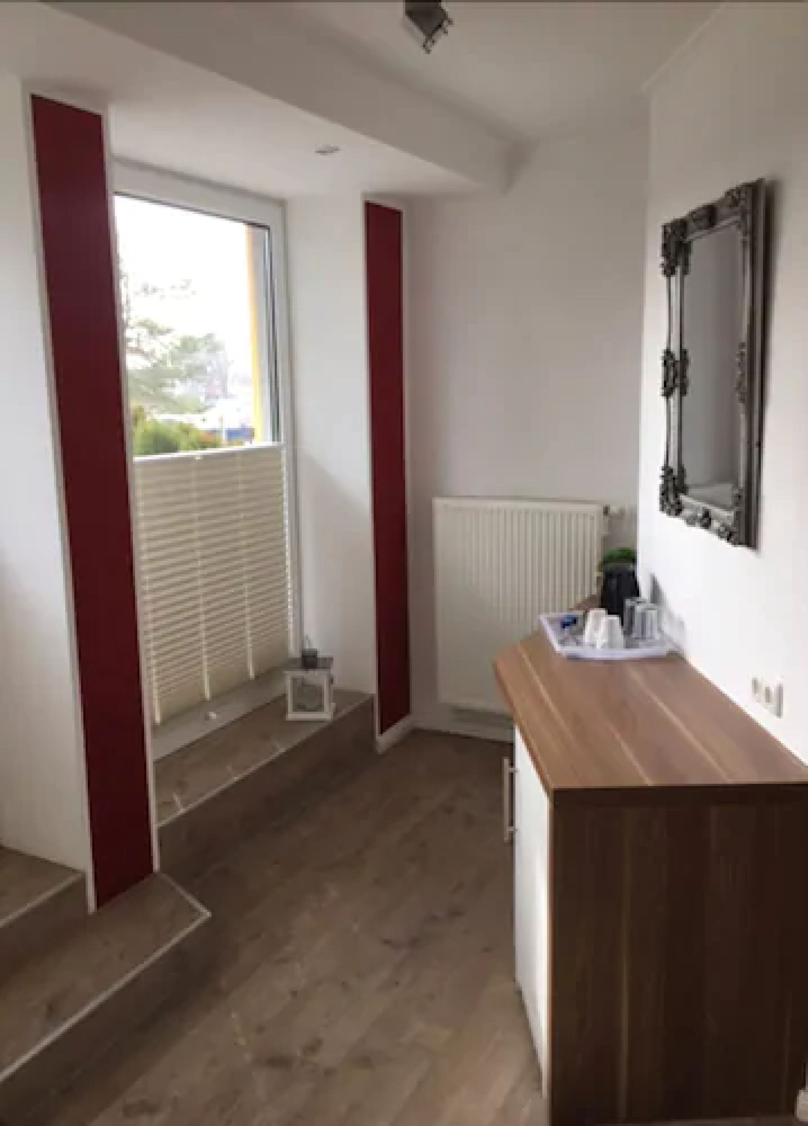 A room with a wooden floor, a radiator, a small table with cups and a bottle, and a window with blinds.