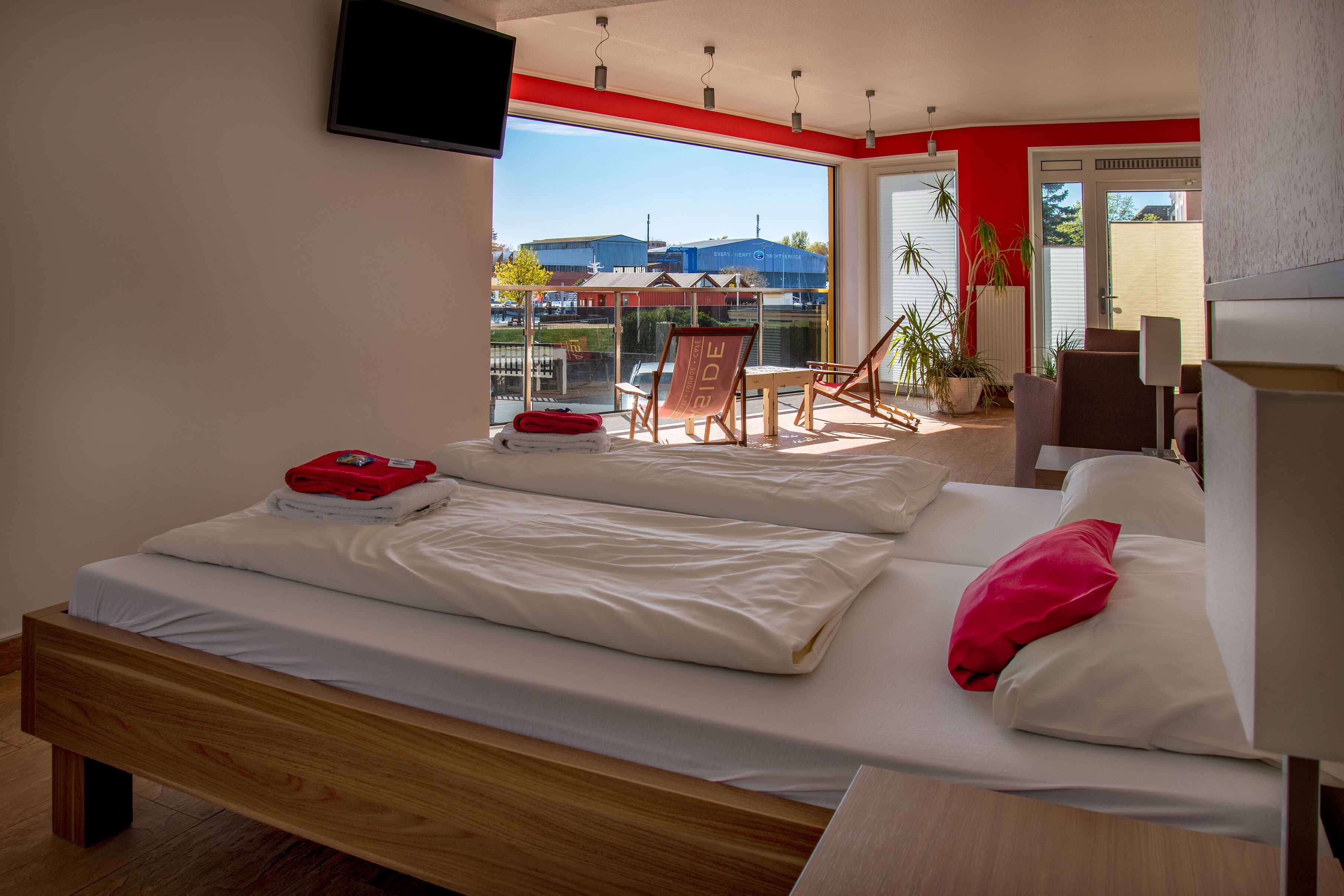 A bedroom with two beds, a balcony with a view of buildings, a TV mounted on the wall, and a chair.