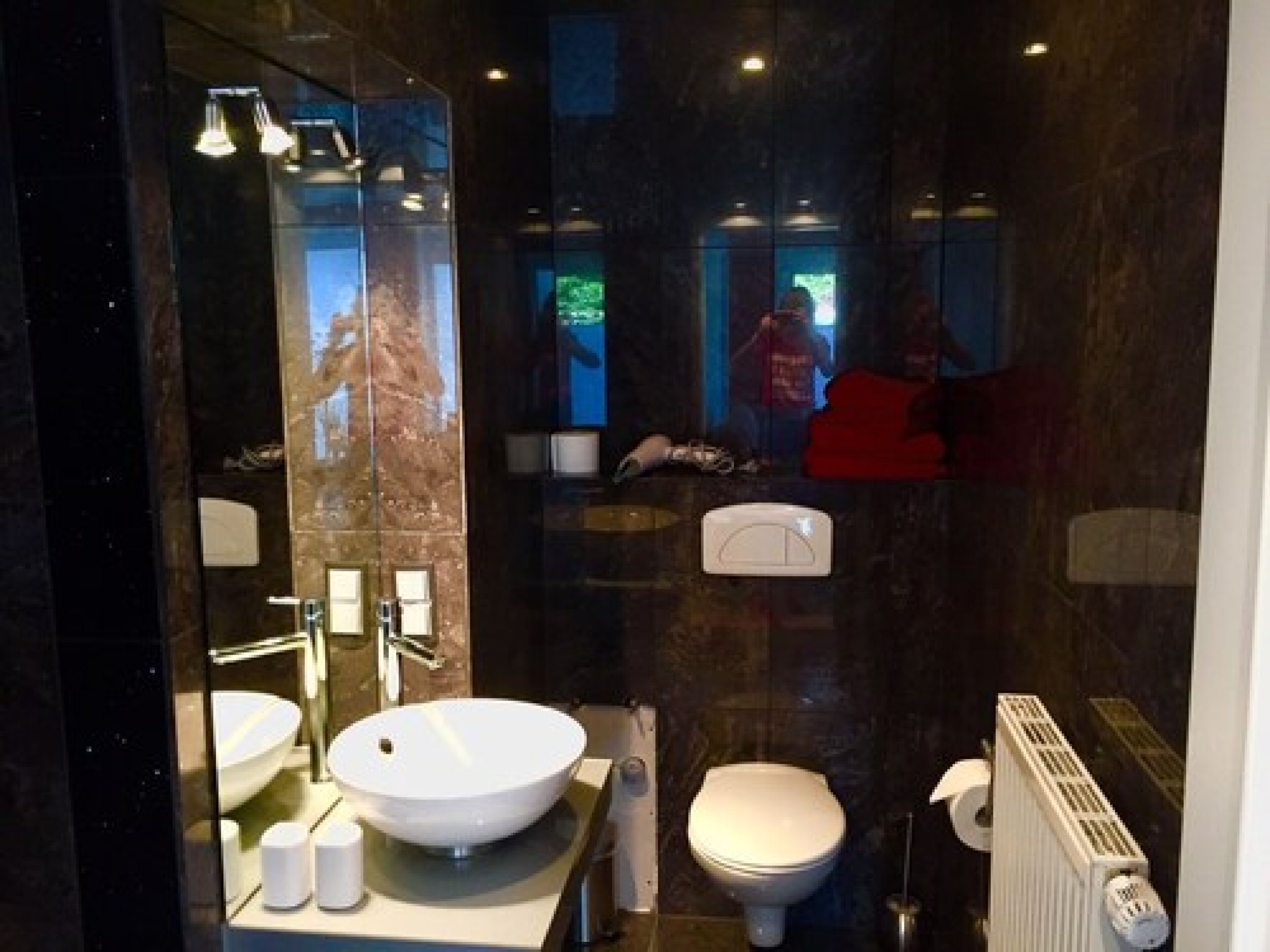 A modern bathroom with a large mirror, a round sink, a toilet, and a radiator.