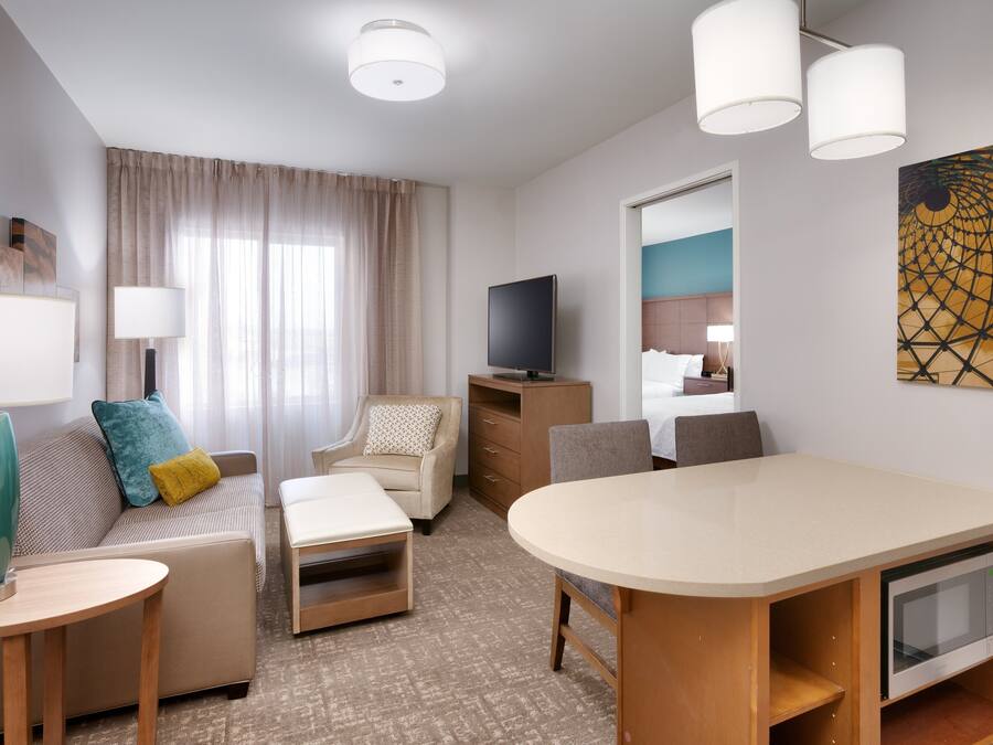 Quarto de hotel com cama, televisão, escrivaninha, cadeira, sofá, abajur e um quadro na parede.