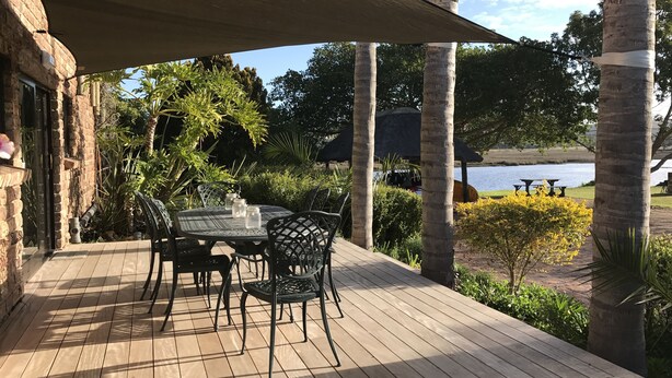 Eine Holzterrasse mit Tisch und Stühlen, umgeben von Palmen und Blick auf einen See.