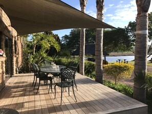 A wooden deck with a table and chairs, surrounded by palm trees and a view of a lake. - Moonriver Self-catering (Plettenberg Bay)