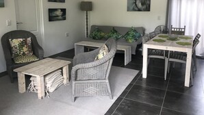 Ein Wohnzimmer mit einer Couch, zwei Sesseln, einem Couchtisch und einem Esstisch mit Stühlen. An den Wänden befinden sich Kunstwerke und es steht eine Lampe bereit.