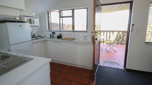 Eine kompakte Küche mit weißen Schränken, einer Mikrowelle, einem Toaster und einem Brothalter. Ein Fenster mit Jalousien und eine Tür zu einem Außenbereich mit einem Stuhl sind vorhanden.