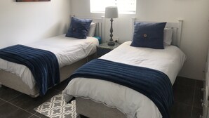 Ein Zimmer mit zwei Einzelbetten, jeweils mit blauem Kopfkissen und weißer Bettwäsche. Es gibt ein Fenster mit Jalousien, einen Nachttisch mit Lampe und ein Bild an der Wand.