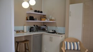 A compact kitchen with a microwave, toaster, and small refrigerator. There is a wicker chair with a cushion and a stool.