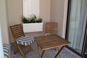 Un petit balcon avec une table en bois et deux chaises à rayures, un pot de fleurs blanc et une porte vitrée avec des rideaux.