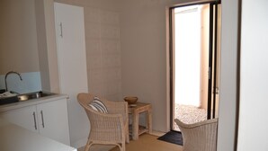 A compact kitchen with a sink, cabinets, and a wicker chair. There's a small table with a bowl and a mat leading to an outdoor area.