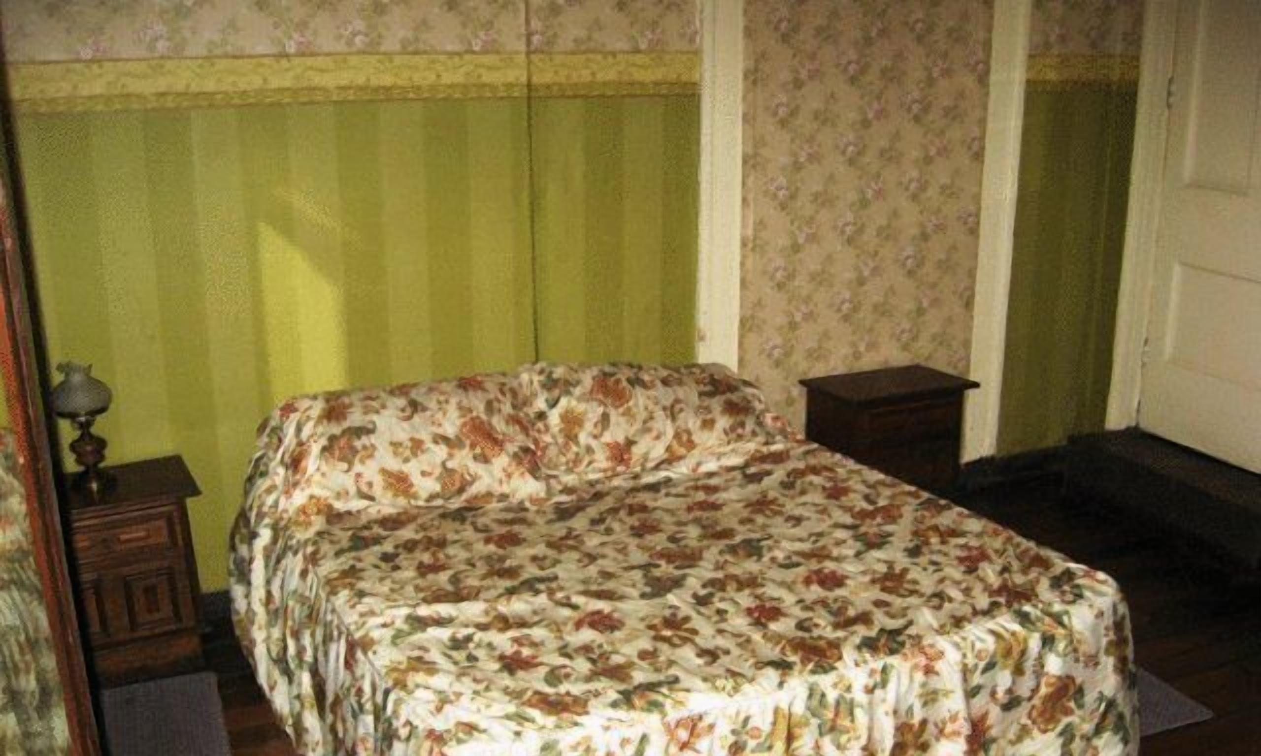 A bedroom with a floral bedspread, a wooden nightstand, and a door with a floral-patterned curtain.