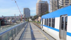 A train station platform with a blue and white train, a pedestrian walkway, and tall buildings in the background. - Hotel Garden (Valparaiso)