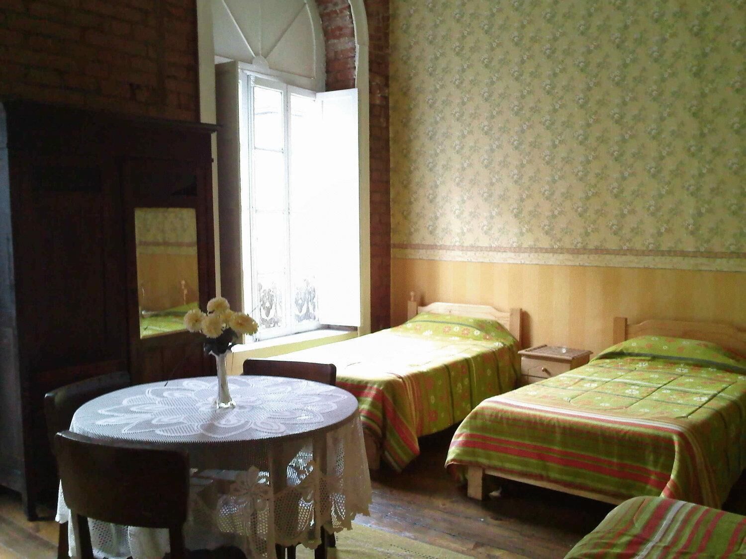 A room with two beds, a table with a lace tablecloth, and a window with a view.