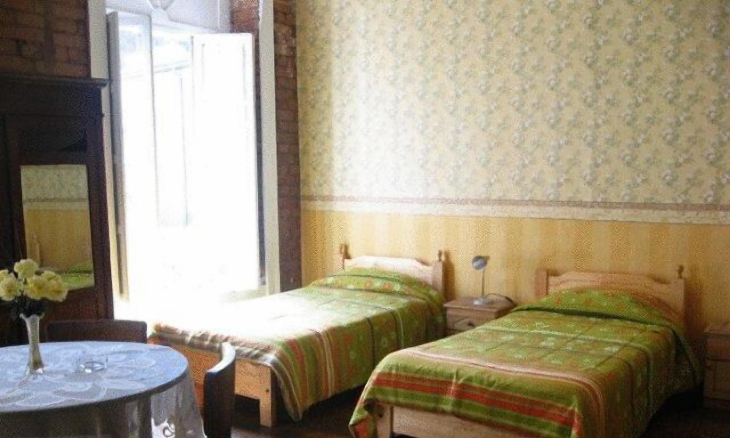 A room with two single beds, a round table with a vase of flowers, and a window with a view of the outside.