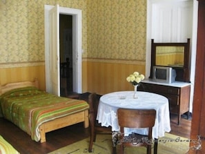 Una habitación con una cama, una mesita con un jarrón de flores, una silla, un escritorio con televisor y un espejo.