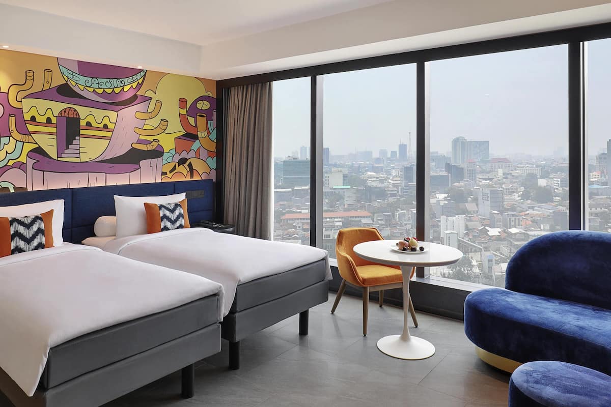 Habitación de hotel con dos camas, una mesita redonda pequeña, un sillón azul y un ventanal con vistas a la ciudad.