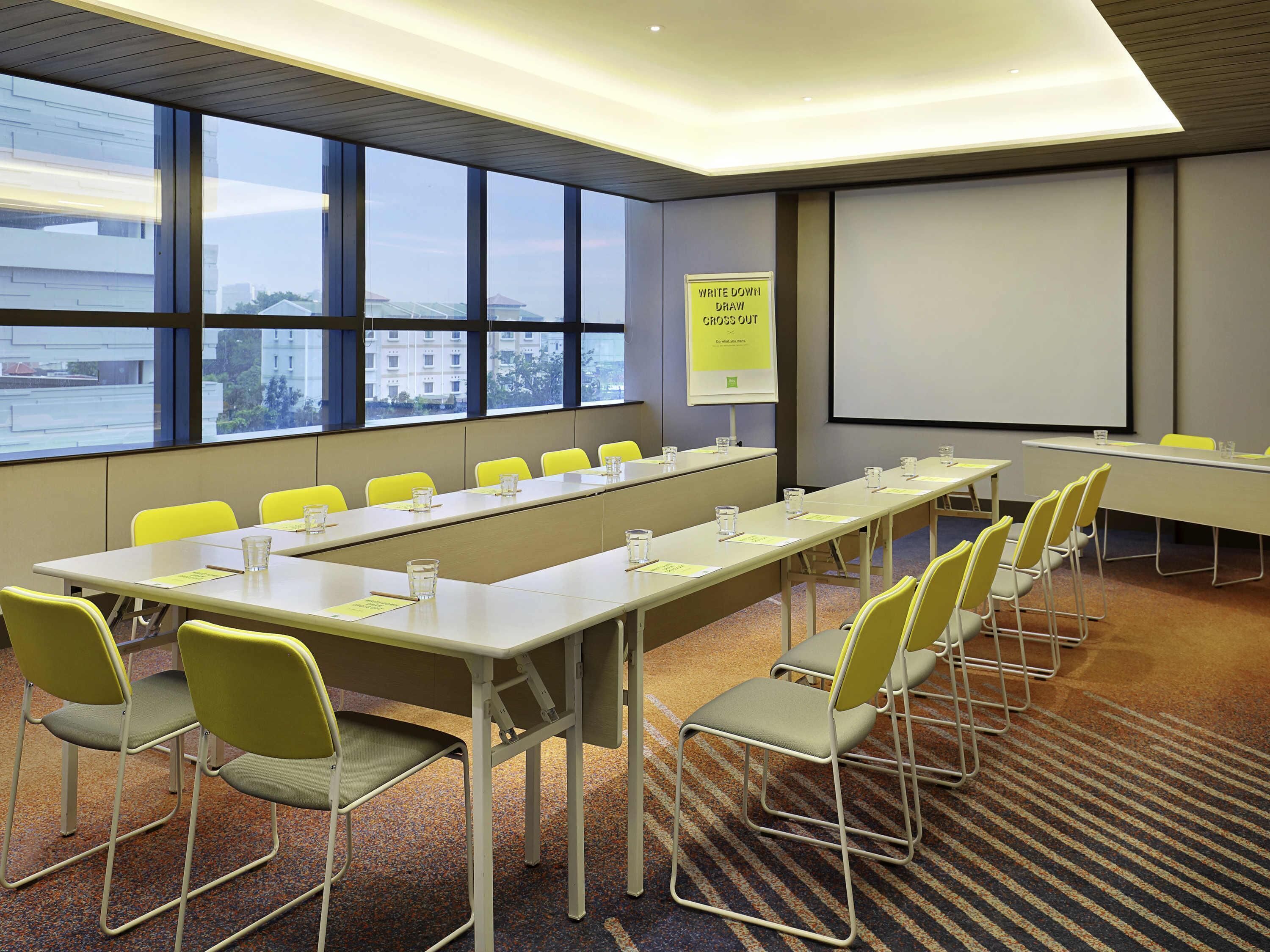 A conference room with a long table, yellow chairs, a projector screen, and a window with a city view.