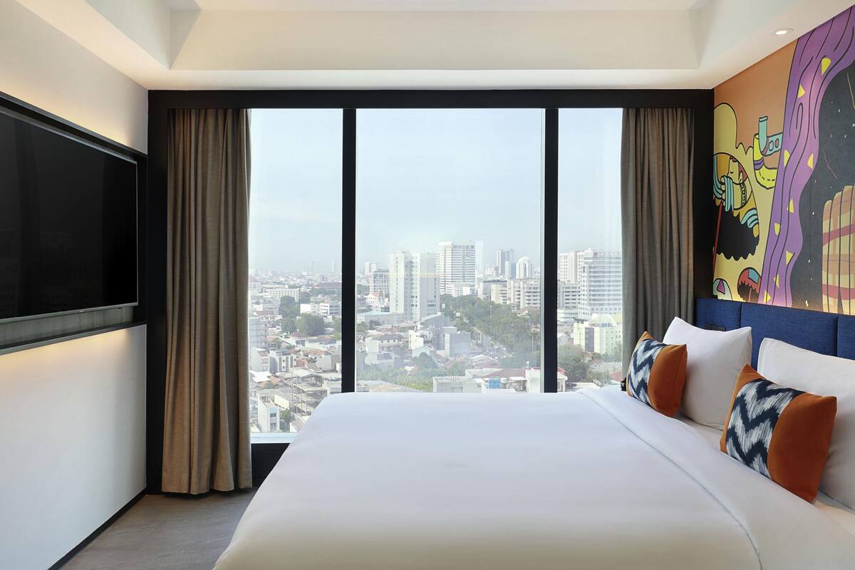 Habitación de hotel con una cama grande, televisión de pantalla plana y vistas a la ciudad a través de ventanales de piso a techo.