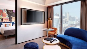 Ein modernes Hotelzimmer mit großem Fernseher, einem Bett, einem blauen Sofa, einem kleinen Tisch mit Laptop und Spielecontrollern sowie Stadtblick durch große Fenster.