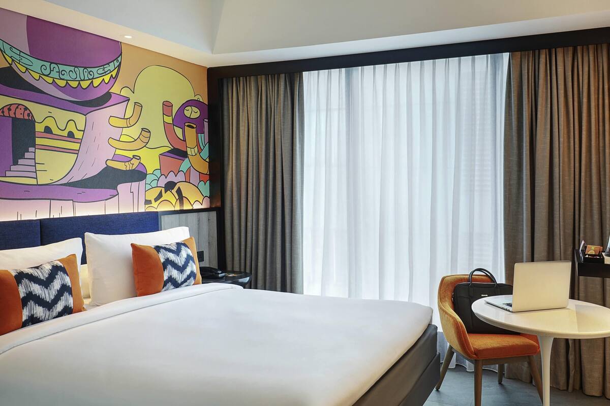 Una habitación de hotel con una cama grande, un escritorio con silla, un cuadro en la pared y una ventana con cortinas.