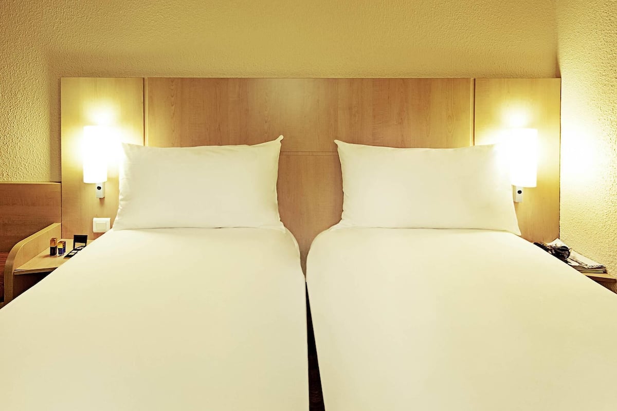 A hotel room with two beds, wooden headboard, bedside lamps, and a nightstand with a phone.