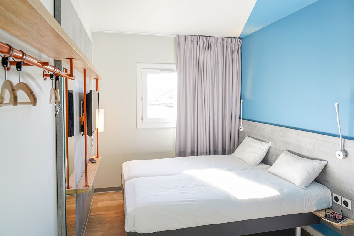 Une chambre à coucher moderne avec un lit, une étagère en bois sur laquelle sont posés divers objets, une fenêtre avec des rideaux et un mur d’accent bleu.
