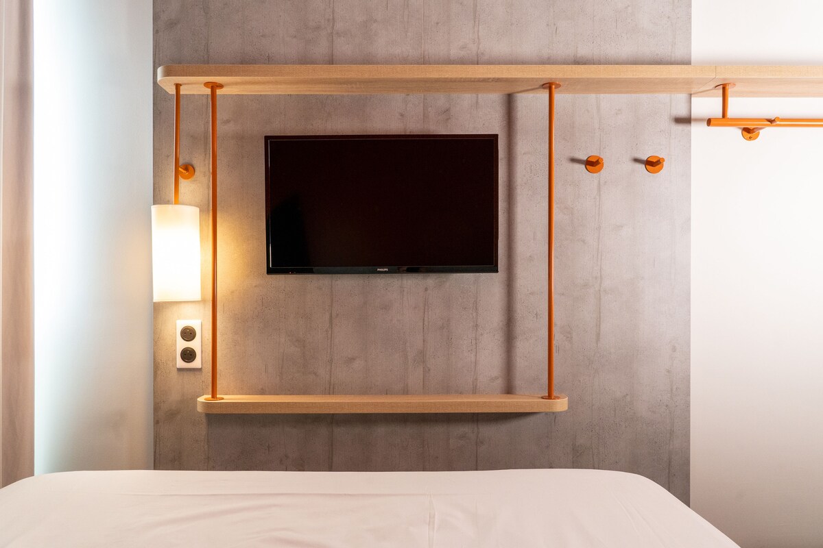 A modern hotel room with a mounted TV, wooden shelf, and wall-mounted lamp.
