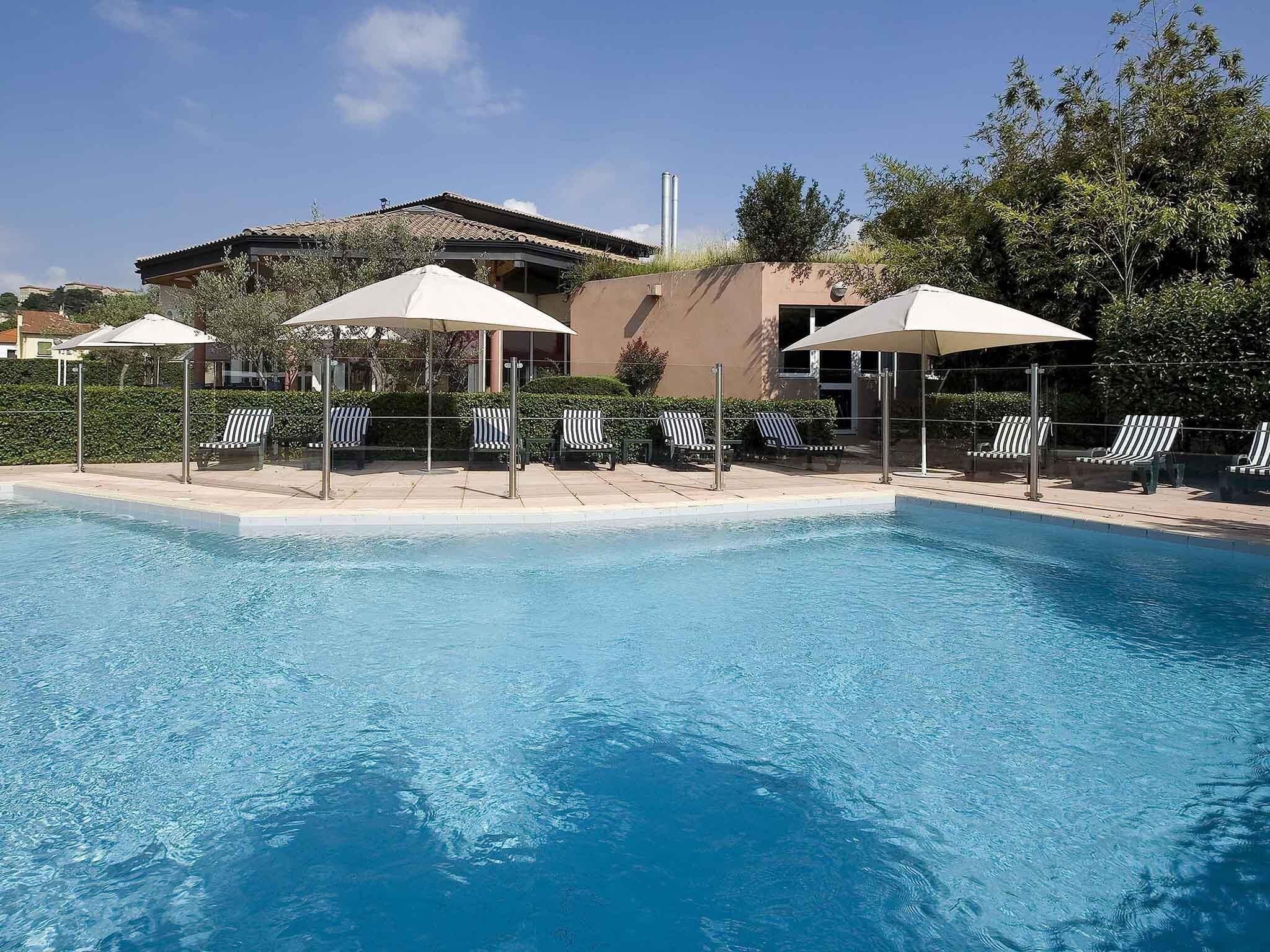 A swimming pool with lounge chairs and umbrellas, surrounded by greenery and a building with a tiled roof.
