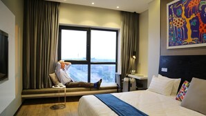 A hotel room with a bed, a window with curtains, a desk, and a painting on the wall. - Room50two (Gaborone)